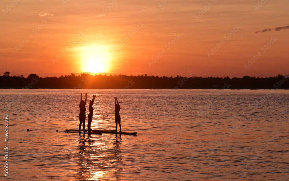 Silhouette of three girls doing yoga trainer balancing on the water on the paddle board over beautiful orange sunset background, Gili Trawangan Island, Indonesia