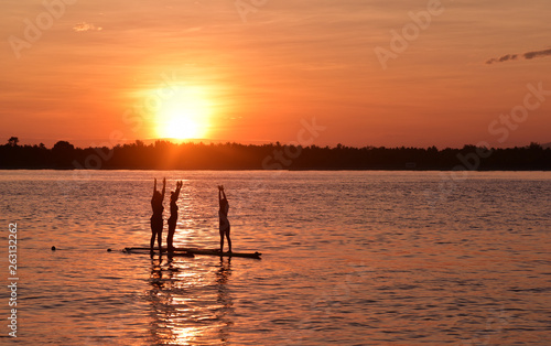 Silhouette of three girls doing yoga trainer balancing on the water on the paddle board over beautiful orange sunset background, Gili Trawangan Island, Indonesia © akturer