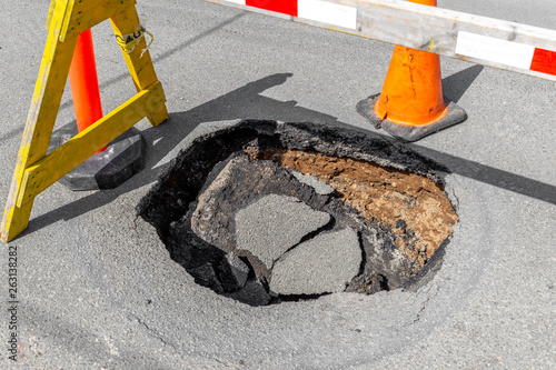 A large, deep pothole in a road. Part of a traffic cone and sawhorse visible blocking the hole. photo