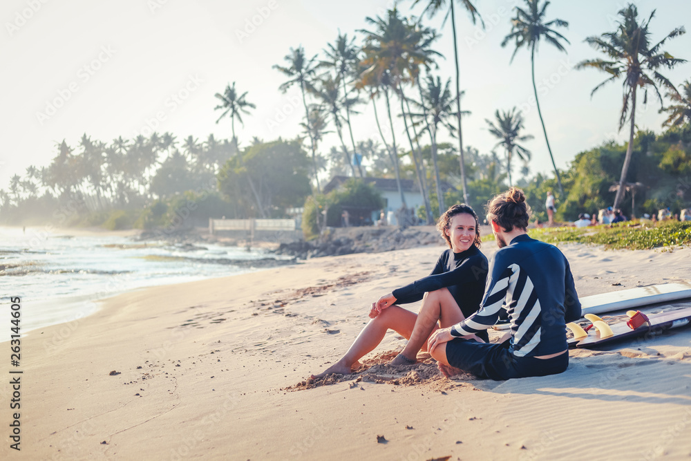 Young beautiful loving couple friends are sitting on the sand by the ocean with surfboards and watching sports active lifestyle holidays honeymoon...