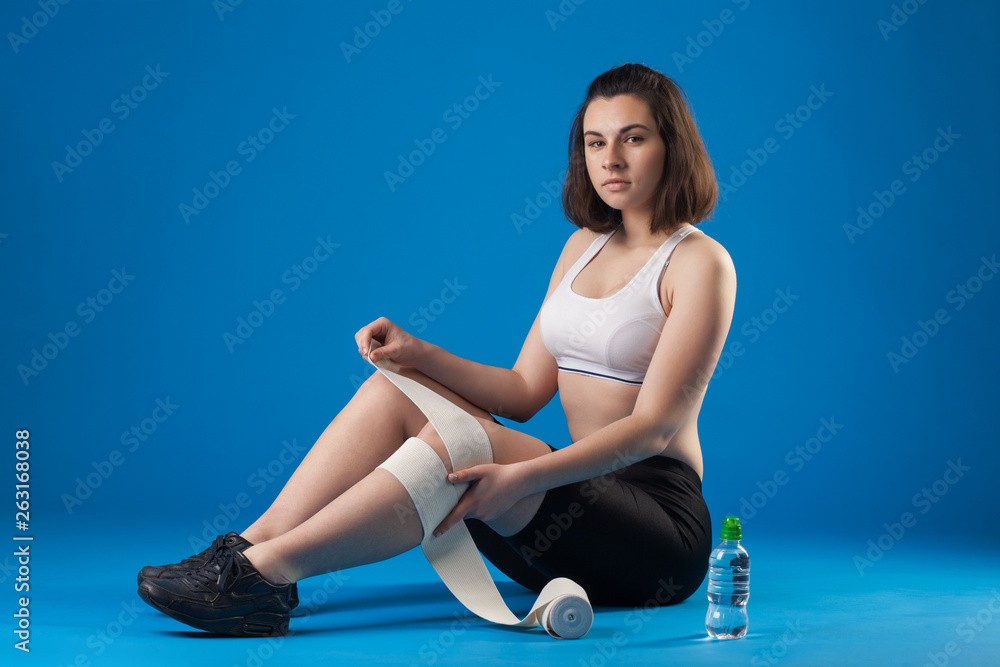 Brunette athlete girl wraps leg with a bandage on a blue background. Sports injury. Brunette athlete girl wraps leg with a bandage on a blue background. Sports injury.