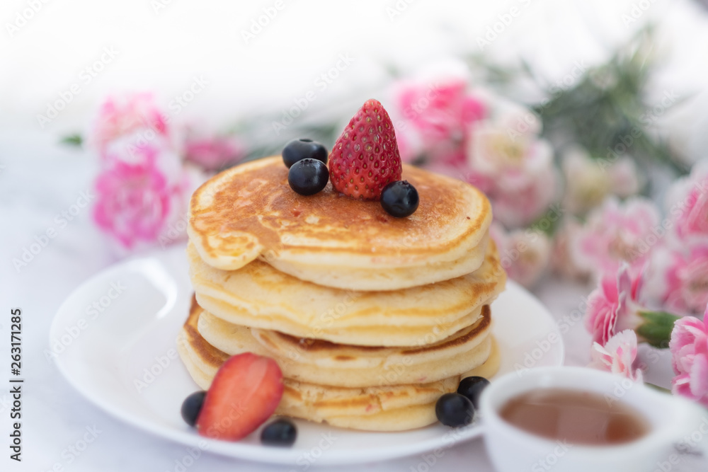 Front view of Pancakes on white plate topped with strawberry, rasberry and black currants and honey syrup and pink blooming flower in blackground.