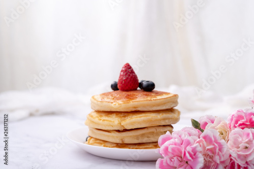 Pancakes on white plate topped with strawberry, rasberry and black currants and honey syrup and pink blooming flower in blackground.