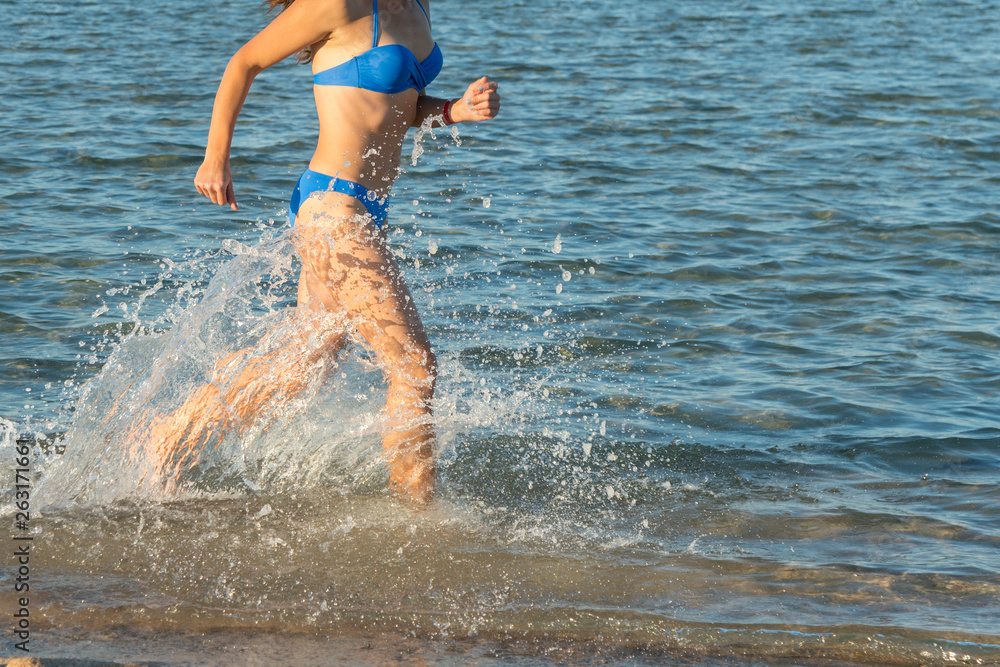 A sexy young brunette woman or girl wearing a bikini running through the surf on a deserted tropical beach with a blue sky. Young woman running by the sea