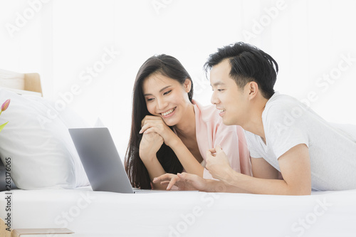 Asian couple lie prone on bed in bedroom.