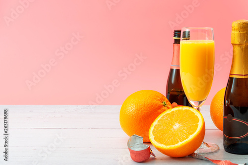 Lush mix drink, summer alcoholic beverages and refreshing mimosa cocktail concept with oranges, champagne flute and bottles of bubbly isolated on white wooden table and pink background with copy space