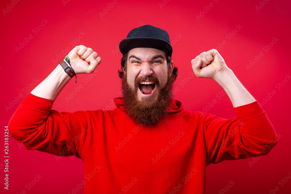 Photo of handsome bearded man in red hoody, looking at camera while doing a victory gesture with rised arms Photo of handsome bearded man in red hoody, looking at camera while doing a victory gesture with rised arms