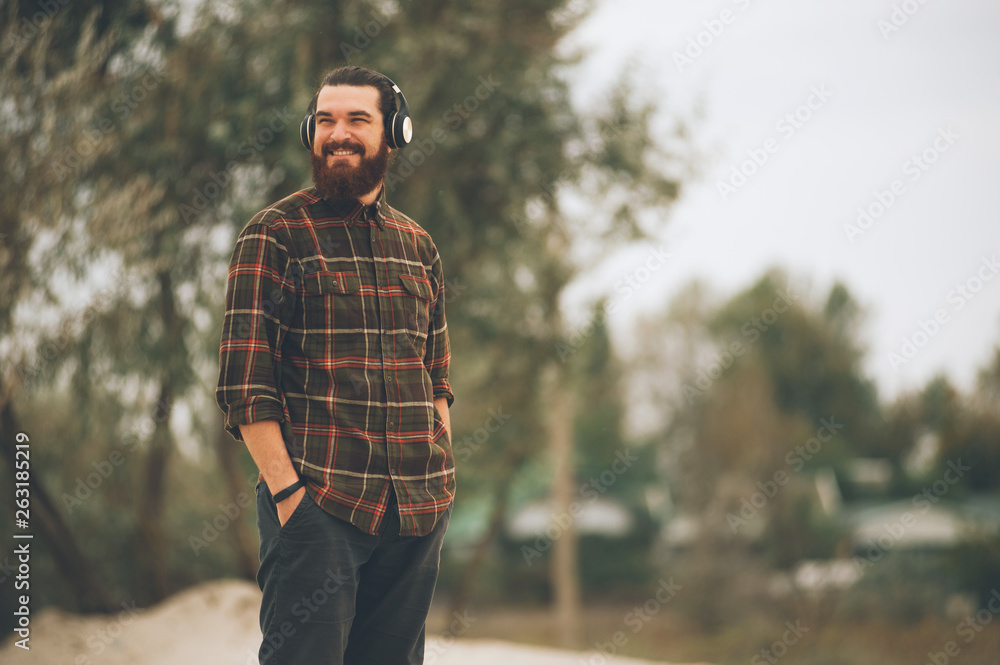 Photo of a smiling bearded man listening music with headphones, putting hands in pockets, and looking  far away