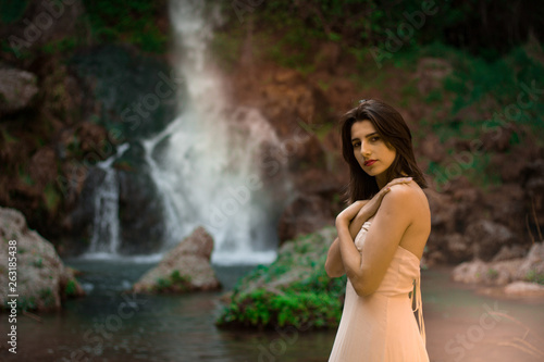 Sexy woman posing on the rock among green tropical plants and beside beautiful waterfall with blue water. Fairy tale.