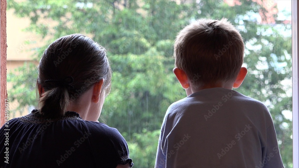 Mother and son admire summer rain at window, boy touching drops Mother and son admire summer rain at window, boy touching drops