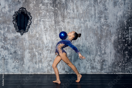 Fototapeta Naklejka Na Ścianę i Meble -  Girl gymnast in a blue suit makes exercise with a ball against a gray wall.