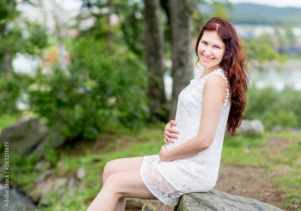 Portrait of cute young caucasian woman in nice white dress in the summer park. Pregnant model sits on a stone and holds her stomach.