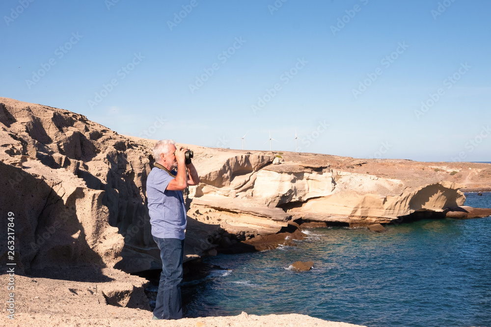 senior man standing with a binoculars on a rock and looking at the sea
