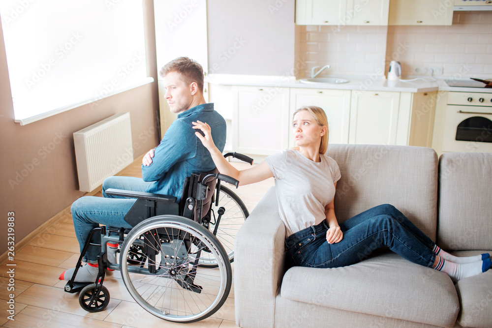 Young man with special needs sit on wheelchair back to back with woman. She touch his shoulder with hand and look at him. Upset and unhappy couple. Young man with special needs sit on wheelchair back to back with woman. She touch his shoulder with hand and look at him. Upset and unhappy couple.