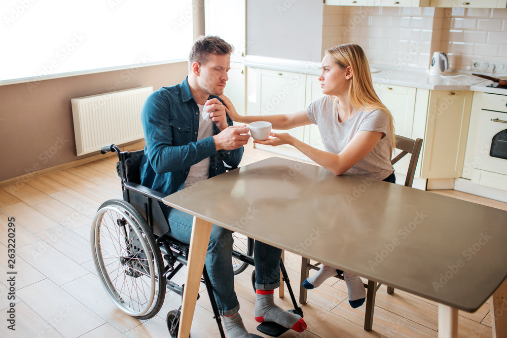 Young woman take care of man with special needs. He sit on wheelchair and get cup of hot drink. Sick and ill. Man with disability and inclusiveness.