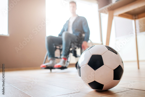 Young man with disability sitting on wheelchair and look down at ball for game. Ex sportsman. Upset and unhappy. Trauma. Can't play football anymore. photo