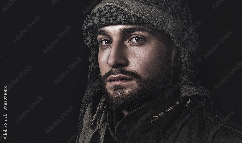 young man with beard in arabic headscarf and military jacket on black background