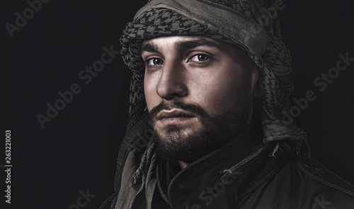 young man with beard in arabic headscarf and military jacket on black background