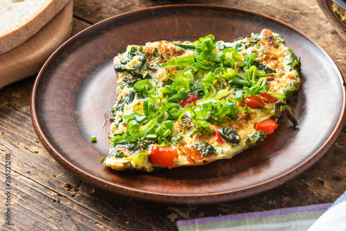 Home-made frittata with spinach and red pepper, sprinkled with spring onions on a plate and cutting board with bread, rustic style, close-up, top view