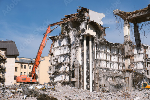 Building demolition with hydraulic excavator. Dismantle of destructed house ruins at bright sunny day with clear blue sky. © Arsenii