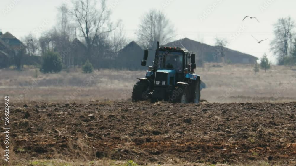 Static shot of a blue tractor with big black wheels and a powerful grouser, heavy plowing the dark fertile soil. The farm machine is followed by white birds. The warmth from the ground and the motor Static shot of a blue tractor with big black wheels and a powerful grouser, heavy plowing the dark fertile soil. The farm machine is followed by white birds. The warmth from the ground and the motor