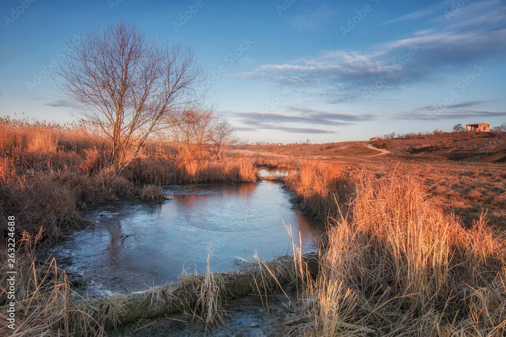 Spring morning on the river bank covered with a thin crust of ice. In the blue sky fluffy clouds. Rural landscape.