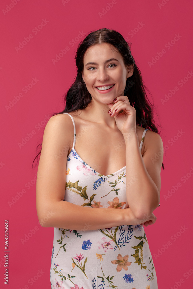 Portrait of a pretty young woman in a light dress standing on pink background in studio. People sincere emotions.