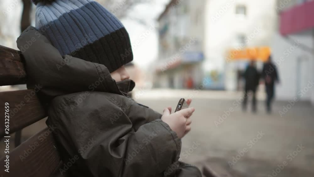 Little boy addictive smartphone sitting on bench in city street. Cute baby boy child with mobile phone on bench outdoor. Little boy addictive smartphone sitting on bench in city street. Cute baby boy child with mobile phone on bench outdoor.