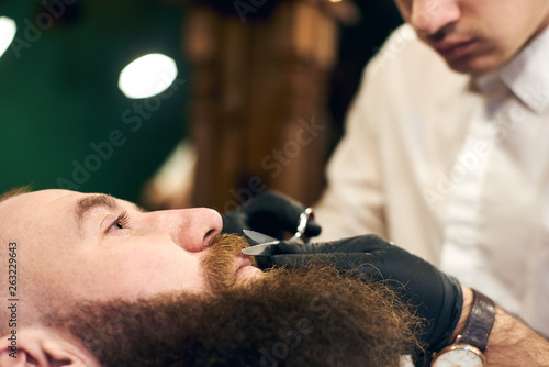Cutting mustache with sharp scissors. Barber in black hygienic gloves concentrate in scrupulous work process. Mustache styling and cutting. Advertising for barber shop. Side view. Blurred background.