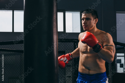 Muscular shirtless athlete in red boxing gloves throwing punch in dark boxing gym. Confident boxer with tattoo on arm doing boxing cardio hitting punch bag. Boxing practice.
