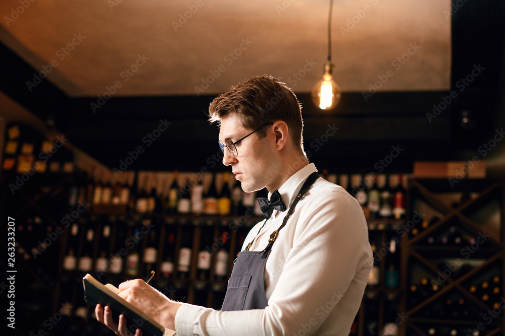 Professional male cavist taking notes at degustation card standing near wine shelves of european wine boutiuque