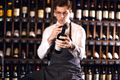 Young caucasian cavist dressed in white shirt and bowtie working in big vine shop presenting a bottle of red wine to customer photo