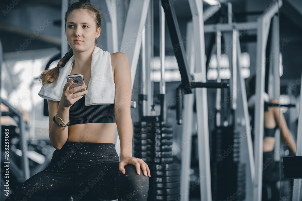 Fototapeta premium Szczęśliwy młodych atrakcyjnych kobiet fitness za pomocą smartfona odpoczywa w siłowni podczas porannych ćwiczeń. Relaks po ciężkim treningu na siłowni. Zdrowy styl życia sportowego, koncepcja fitness.