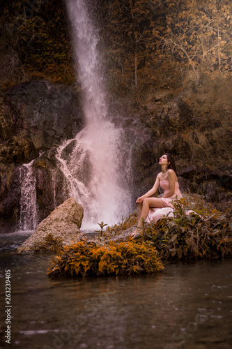 Attractive girl posing on the rock among in fairy fast forest in autumn and beside beautiful waterfall with blue water. Fairy tale.