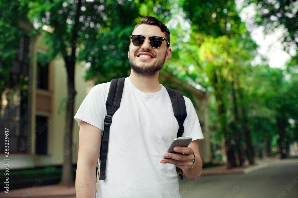 Stylish unshaven hipster wearing white tshirt, black bagpack and sunglasses using a smart phone outdoor Stylish unshaven hipster wearing white tshirt, black bagpack and sunglasses using a smart phone outdoor