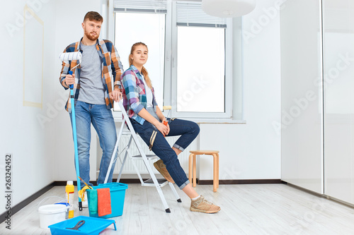 Portrait of smiling couple painting at home, holding paint roller and brush, looking at camera.