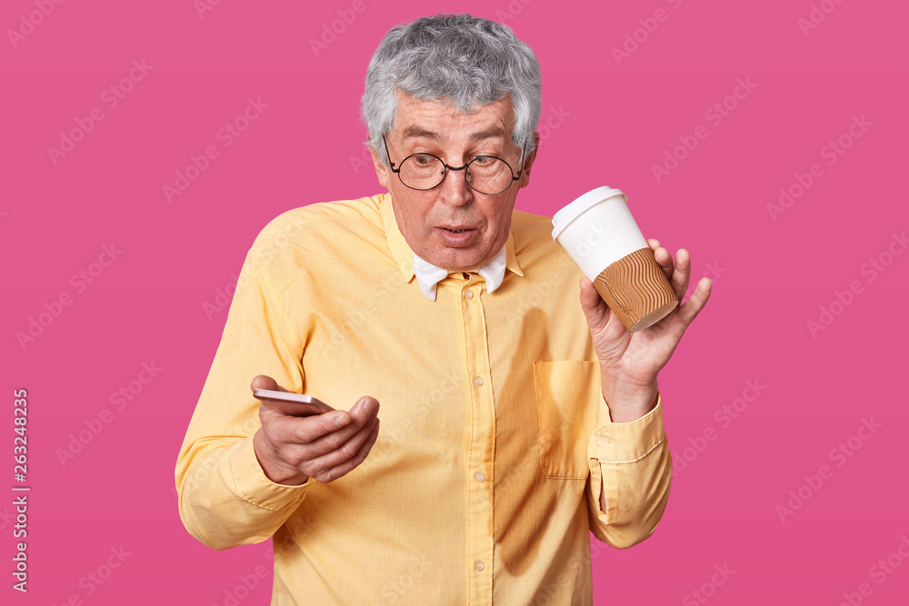Astonished old man poured coffe on his yellow shirt while reading shocking information in his smart phone, wears glasses, has surprised facial expression, poses over pink studio wall, empty space. Astonished old man poured coffe on his yellow shirt while reading shocking information in his smart phone, wears glasses, has surprised facial expression, poses over pink studio wall, empty space.