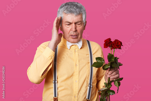Studio shot of grey haired elderly man in fashionable yellow shirt, suspenders, white bow tie, keeps hand on temple, foegets about something important, holds red roses, poses over pink background. photo