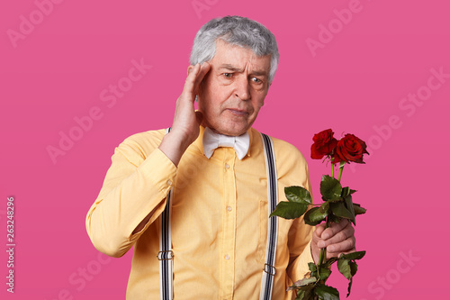 Image of romantic mature gentleman in yellow shirt with bowtie and suspenders, holds red roses, has problems, going for date or birthday, keeps hand on head, looks aside with confused expression. photo