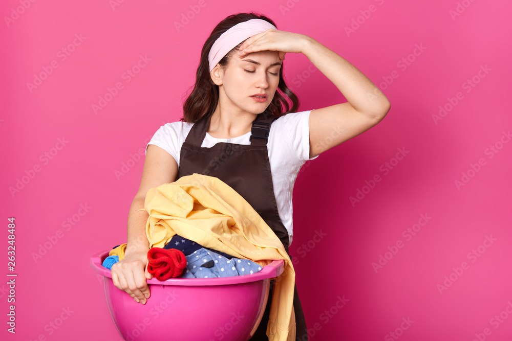 Slender stressful young female puts her hand on head, holding basin with dirty clothes, closing her eyes, being exhausted of house work, turning her head aside. Household and domestic concept. Slender stressful young female puts her hand on head, holding basin with dirty clothes, closing her eyes, being exhausted of house work, turning her head aside. Household and domestic concept.