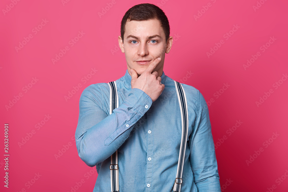 Peaceful thoughtful young boy stands wearing blue shirt, bow tie and stripped suspenders, posing isolated over pink background in studio, looks interested in something. People and emotions concept. Peaceful thoughtful young boy stands wearing blue shirt, bow tie and stripped suspenders, posing isolated over pink background in studio, looks interested in something. People and emotions concept.