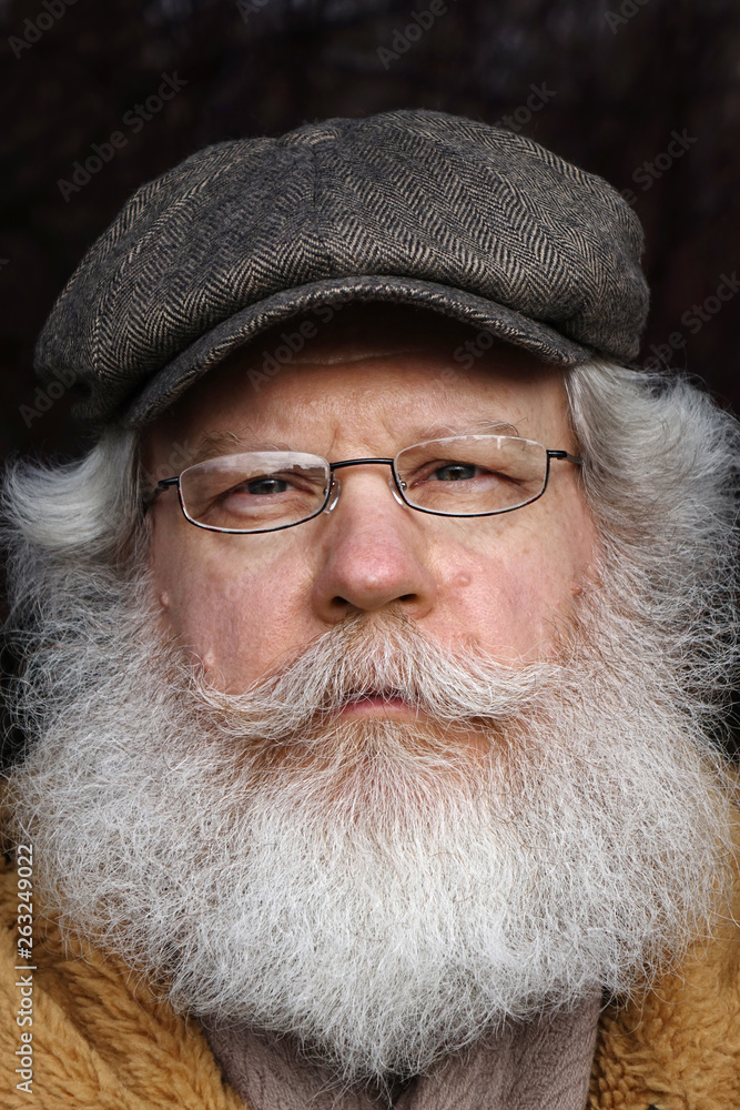 Senior caucasian man looking at camera against dark natural background. Extreme close up vertical face portrait of elderly stylish hipster wearing eyeglasses and kepi with splendid mustache and beard. Senior caucasian man looking at camera against dark natural background. Extreme close up vertical face portrait of elderly stylish hipster wearing eyeglasses and kepi with splendid mustache and beard.