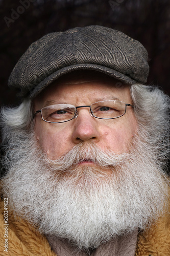 Senior caucasian man looking at camera against dark natural background. Extreme close up vertical face portrait of elderly stylish hipster wearing eyeglasses and kepi with splendid mustache and beard.