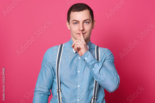 Mysterious handsome model puts his forefinger in front of mouth, showing shh sign, keeps in silent, looks curious. Brunette magnetic European boy stands in studio, looking directly at camera. photo