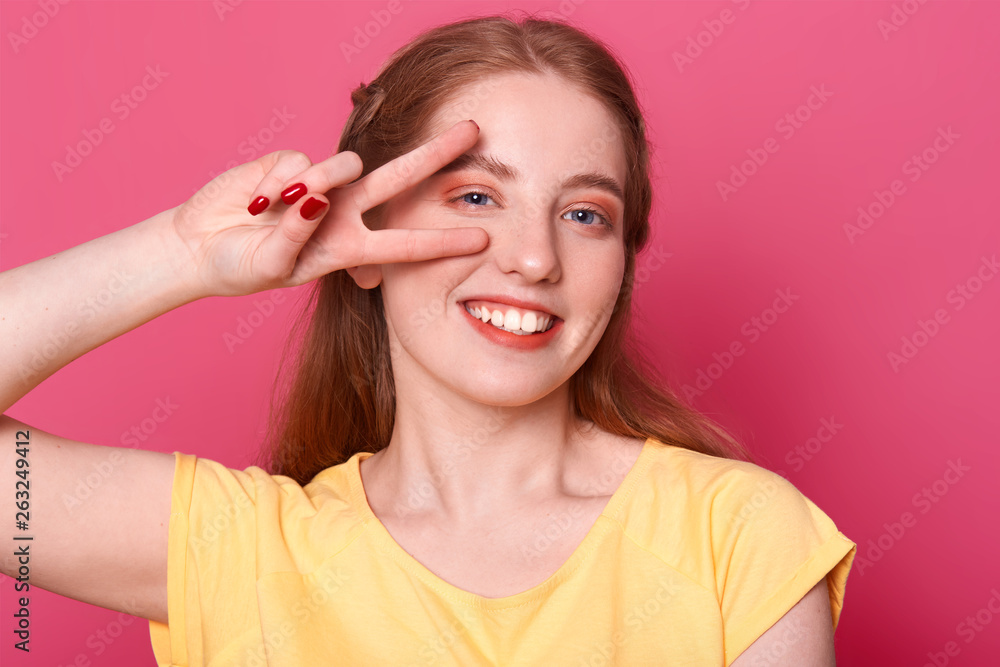 Smiling positive model poses isolated over bright pink background in studio with victory hand near her right eye, wearing yellow tshirt, having pleasant facial expression. Free time and youth concept. Smiling positive model poses isolated over bright pink background in studio with victory hand near her right eye, wearing yellow tshirt, having pleasant facial expression. Free time and youth concept.