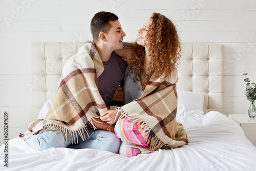 Fototapeta Photo of girlfriend and boyfriend dressed casually, enjoy coziness under plaid in bedroom, embrace each other, look into eyes and have pleasant talk
