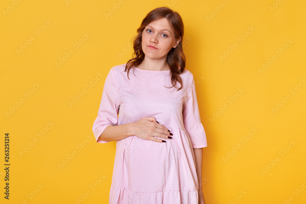 Studio shot of young pretty pregnant woman wearing pink powder dress and holding hand on belly, looking directly at camera with pleasant facial expression, isolated over yellow studio background. Studio shot of young pretty pregnant woman wearing pink powder dress and holding hand on belly, looking directly at camera with pleasant facial expression, isolated over yellow studio background.