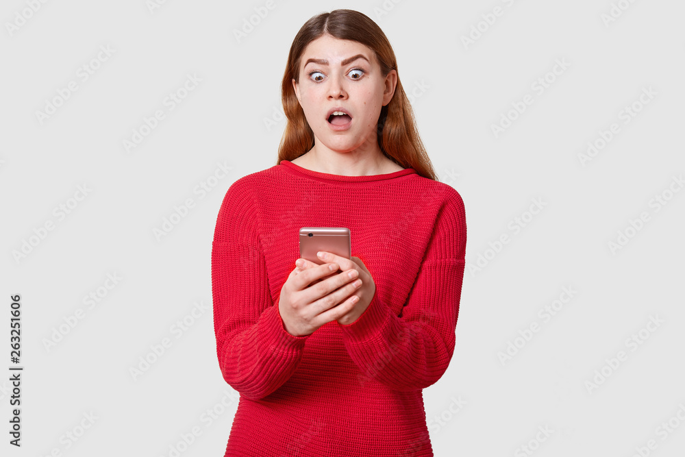 Front view of surprised model holds her smartphone with both hand, looking at display with astonished facial expression, has shoking news fron friend, reading message, isolated over white background. Front view of surprised model holds her smartphone with both hand, looking at display with astonished facial expression, has shoking news fron friend, reading message, isolated over white background.