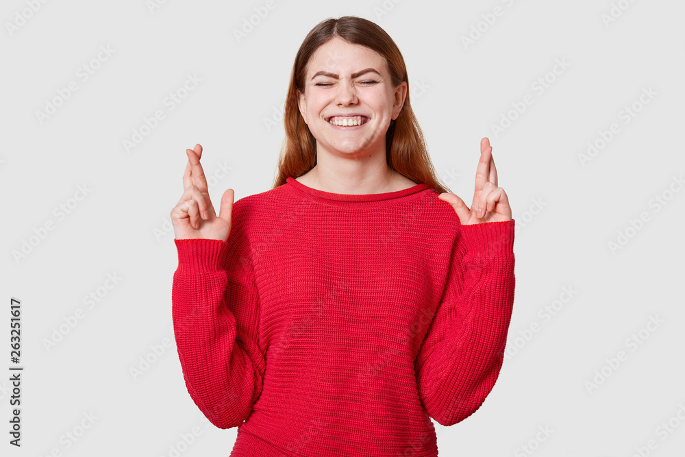 Indoor shot of pleasant looking dressed in casual red sweater, crosses fingers and has great hope for better, waits results of examination, posing in studio with cluosed eyes and toothy smile. Indoor shot of pleasant looking dressed in casual red sweater, crosses fingers and has great hope for better, waits results of examination, posing in studio with cluosed eyes and toothy smile.