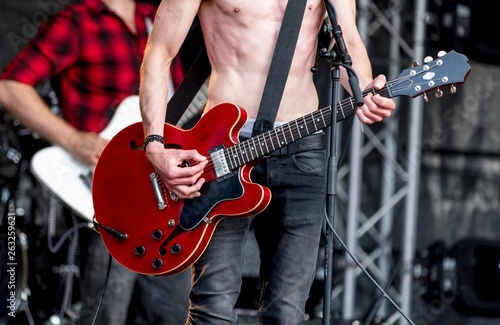 Rock band fronted by heavy metal singer with his top off showing his slim muscular body playing a red electric guitar on stage at a festival